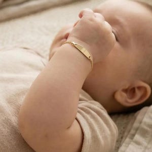 May include: A gold baby ID bracelet with a rectangular nameplate is worn on a baby's wrist. The bracelet is on a baby's arm, and the baby is wearing a beige onesie. The baby is lying on a beige blanket.