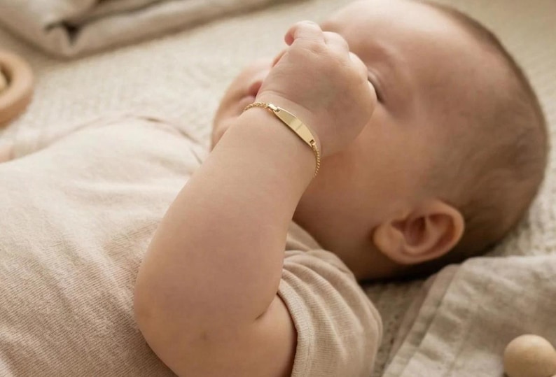May include: A gold baby ID bracelet with a rectangular nameplate is worn on a baby's wrist. The bracelet is on a baby's arm, and the baby is wearing a beige onesie. The baby is lying on a beige blanket.