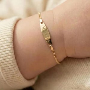 May include: A gold-colored baby bracelet with a nameplate that reads "Mustafa" in a script font. The bracelet is attached to a delicate chain and is worn on a baby's wrist, against a cream-colored sweater.