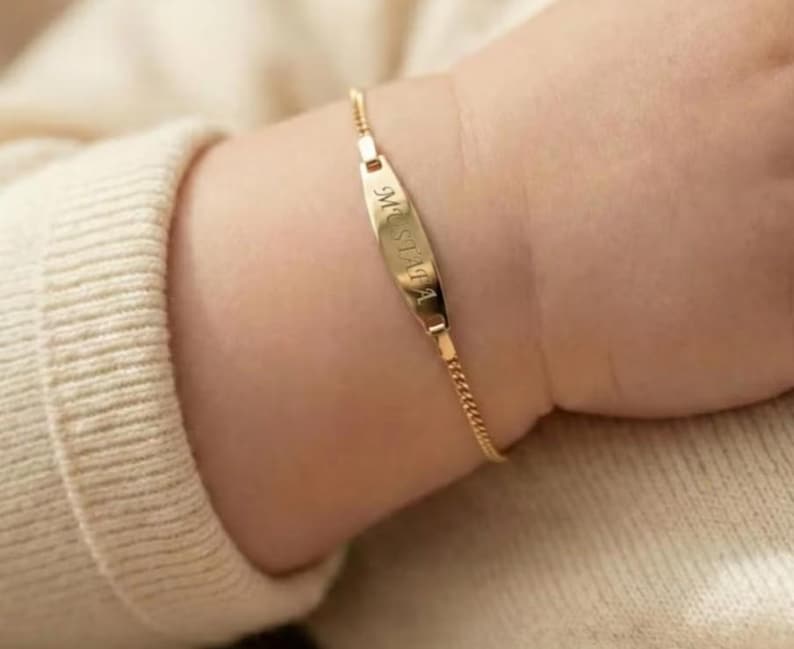 May include: A gold-colored baby bracelet with a nameplate that reads "Mustafa" in a script font. The bracelet is attached to a delicate chain and is worn on a baby's wrist, against a cream-colored sweater.
