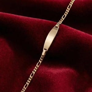 May include: A gold-colored bracelet with a rectangular nameplate and a chain-link design. The bracelet is displayed on a deep red velvet surface, creating a luxurious feel. The bracelet's clasp is visible.