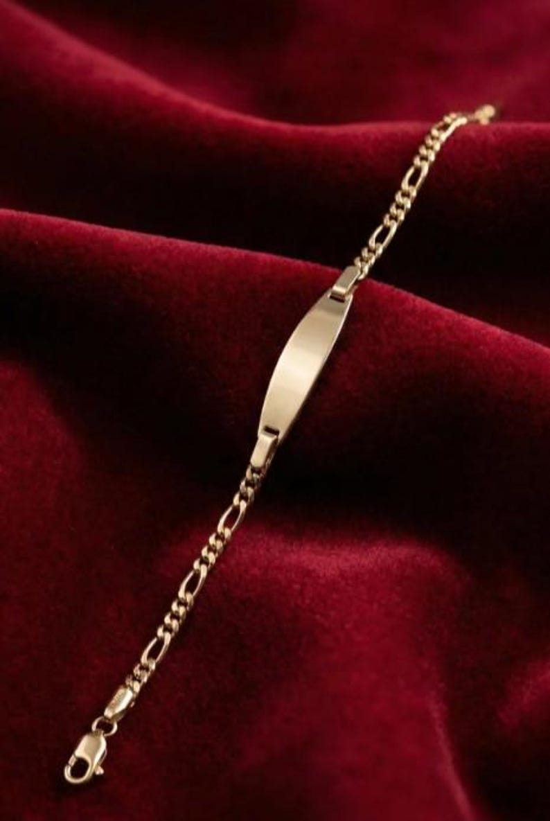 May include: A gold-colored bracelet with a rectangular nameplate and a chain-link design. The bracelet is displayed on a deep red velvet surface, creating a luxurious feel. The bracelet's clasp is visible.