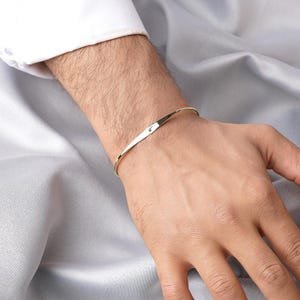 Op de afbeelding: Een goudkleurige armband met een minimalistisch ontwerp. De armband is dun en gebogen, met een klein, subtiel detail aan de voorkant. De afbeelding toont de armband om een pols, tegen een achtergrond van een zijdeachtige, grijze stof.