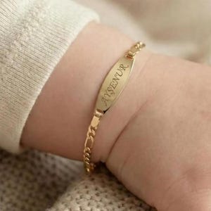 May include: A gold-colored ID bracelet with the name "AYŞENUR" engraved on the oval plate. The bracelet is attached to a small wrist with a gold chain. The background is a soft, neutral color.
