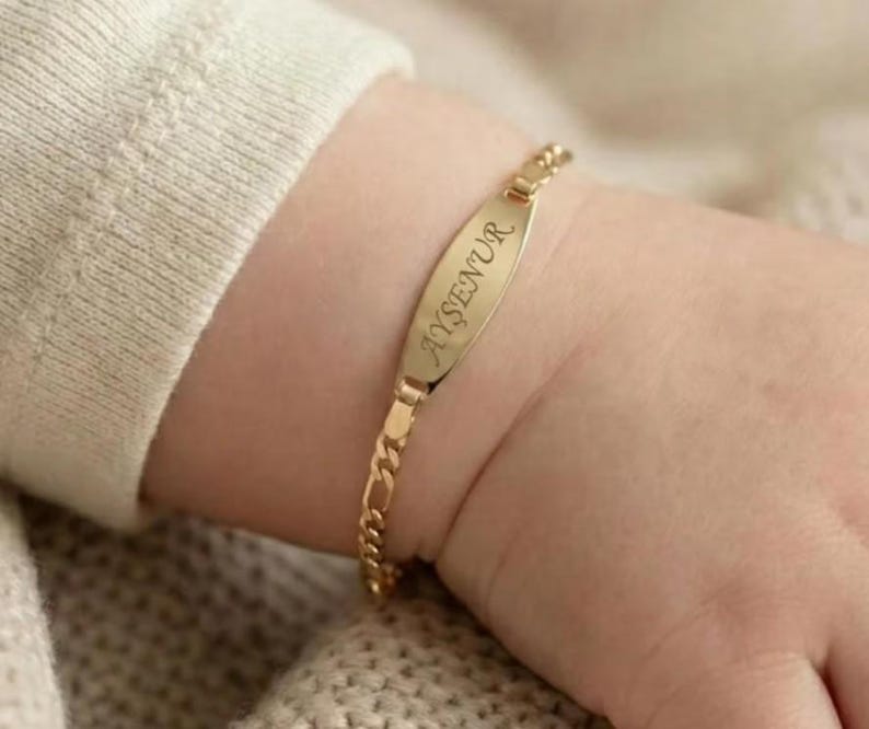 May include: A gold-colored ID bracelet with the name "AYŞENUR" engraved on the oval plate. The bracelet is attached to a small wrist with a gold chain. The background is a soft, neutral color.
