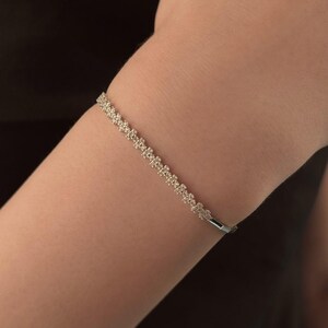 May include: A delicate silver bracelet with a floral design. The bracelet is adorned with small, sparkling stones, creating a shimmering effect. The jewelry is worn on the arm against a dark background.