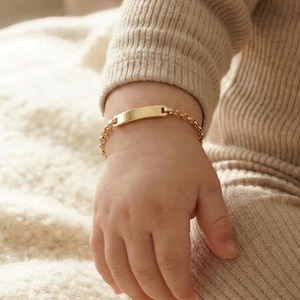 May include: A gold-colored baby ID bracelet with a rectangular nameplate and a delicate chain. The bracelet is shown on a baby's wrist, with the focus on the jewelry. The bracelet is a classic piece of baby jewelry.