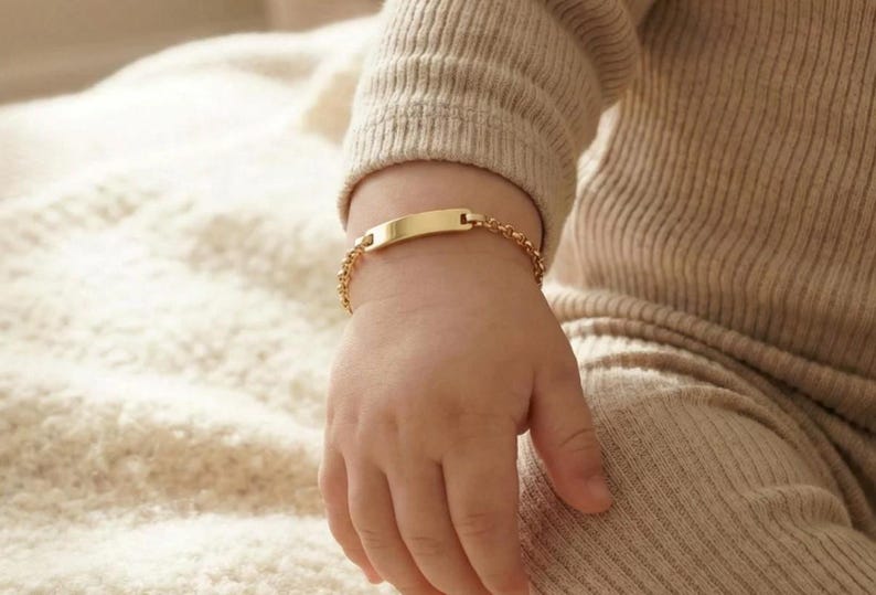 May include: A gold-colored baby ID bracelet with a rectangular nameplate and a delicate chain. The bracelet is shown on a baby's wrist, with the focus on the jewelry. The bracelet is a classic piece of baby jewelry.
