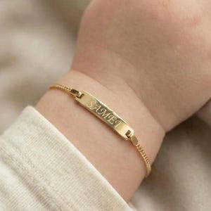May include: A gold-colored baby bracelet with the name "SAMET" engraved on a rectangular plate. The bracelet is attached to a delicate chain and is worn on a baby's wrist.