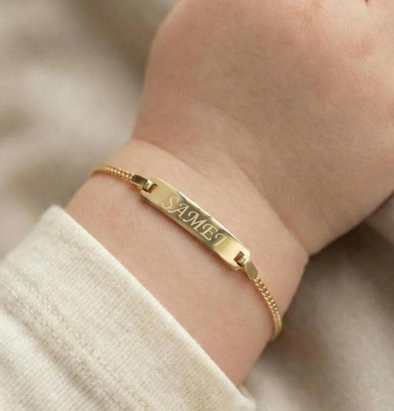May include: A gold-colored baby bracelet with the name "SAMET" engraved on a rectangular plate. The bracelet is attached to a delicate chain and is worn on a baby's wrist.