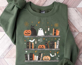 May include: Green sweatshirt with a graphic print of a bookshelf with pumpkins, a black cat, a ghost, and books. The bookshelf is decorated with stars and spiderwebs.
