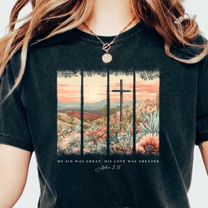 May include: Black t-shirt with a graphic of a cross in a desert landscape. The text "My sin was great, His love was greater John 3:16" is printed below the graphic.