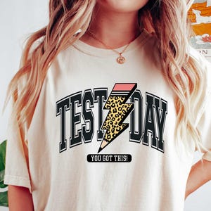 May include: A cream colored t-shirt with a black graphic that says "Test Day" with a pink and leopard print lightning bolt. The text below the graphic says "You got this!"