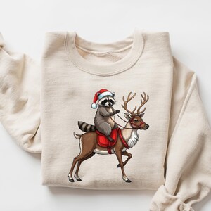 May include: A beige sweatshirt with a graphic of a raccoon wearing a Santa hat riding a reindeer.