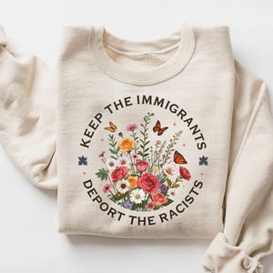 May include: Cream-colored sweatshirt with a floral design and the text "Keep the Immigrants, Deport the Racists." The graphic includes colorful flowers and butterflies. The sweatshirt has long sleeves and a crew neck.