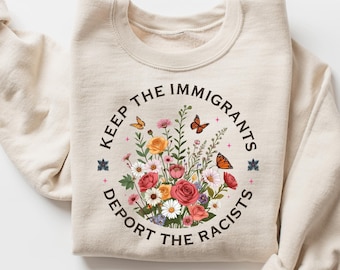 May include: Cream-colored sweatshirt with a floral design and the text "Keep the Immigrants, Deport the Racists." The graphic includes colorful flowers and butterflies. The sweatshirt has long sleeves and a crew neck.