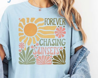 May include: Light blue t-shirt with the text "FOREVER CHASING SUNSETS" in a retro design. The graphic includes a sun, flowers, and foliage in orange, yellow, pink, and green. The shirt is part of a casual wear collection.