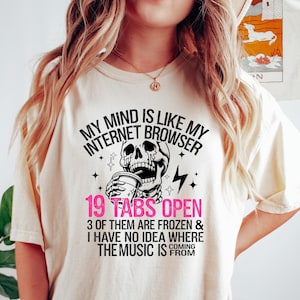 May include: A white t-shirt with a black and white graphic of a skull holding a coffee cup. The text on the shirt reads "My mind is like my internet browser 19 tabs open 3 of them are frozen & I have no idea where the music is from coming".