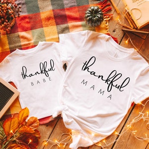 May include: Two white t-shirts with the text "thankful babe" and "thankful mama" in black script. The shirts are laid out on a wooden surface with a plaid blanket, fall leaves, and string lights.