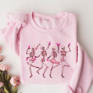 Pode incluir: Camisola rosa com um gr&aacute;fico de quatro esqueletos usando tutus cor-de-rosa e segurando fitas cor-de-rosa. Os esqueletos est&atilde;o dan&ccedil;ando e sorrindo.