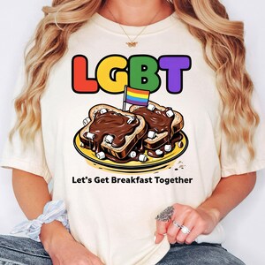 May include: Cream-colored t-shirt with the word "LGBT" in rainbow colors. The shirt features a graphic of two pieces of toast with chocolate and marshmallows, and a rainbow flag. Text below reads "Let's Get Breakfast Together."