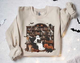 May include: A beige sweatshirt with a graphic print of a ghost reading a book in a library with bats, pumpkins, and black cats. The graphic is in a vintage style.