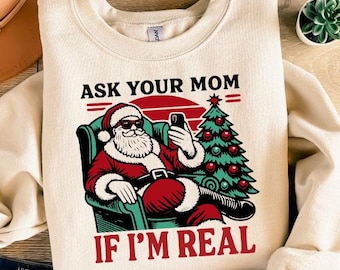 May include: Cream-colored sweatshirt with a graphic of Santa Claus wearing sunglasses, holding a phone, and sitting in a chair. The text reads "ASK YOUR MOM IF I'M REAL". A Christmas tree and a red sun are also in the graphic.