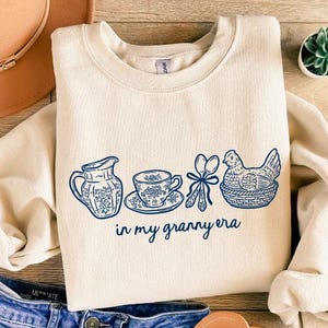 Puede incluir: Sudadera color crema con un dise&ntilde;o floral azul y el texto "in my granny era". El dise&ntilde;o incluye una jarra, una taza de t&eacute;, utensilios y una gallina en una cesta. Un sombrero marr&oacute;n y unos vaqueros azules tambi&eacute;n est&aacute;n en la imagen.
