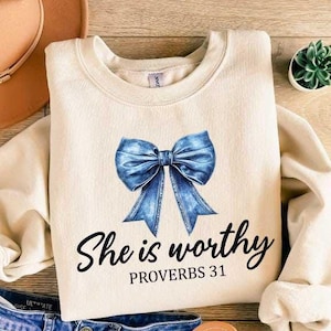 Może przedstawiać: Kremowa bluza z niebieską kokardą z dżinsu i nadrukiem "She is worthy Proverbs 31" z przodu.