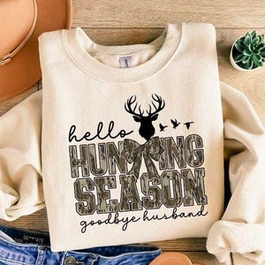 May include: A cream-colored sweatshirt with the text "hello hunting season goodbye husband" in black lettering. The text is surrounded by a camouflage pattern with a silhouette of a deer with antlers and a bow.