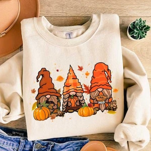 May include: A cream-colored sweatshirt with a graphic of three gnomes wearing orange and brown hats. The gnomes are holding pumpkins and a pie. The sweatshirt has a crew neck and long sleeves.