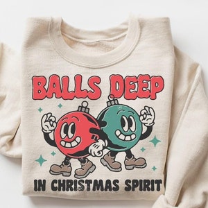 Peut inclure: Un sweat-shirt beige avec un motif de No&euml;l. Le motif repr&eacute;sente deux boules de No&euml;l dessin&eacute;es avec des visages, l'une rouge et l'autre verte, avec le texte "Balls Deep in Christmas Spirit".