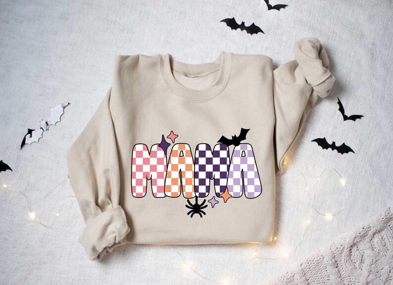 May include: A beige sweatshirt with a checkered "MAMA" graphic in pink, orange, purple, and black. The graphic includes a bat, spider, and stars.