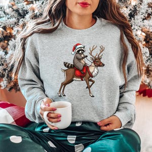 May include: A gray sweatshirt with a Christmas-themed design featuring a raccoon wearing a Santa hat riding a reindeer.