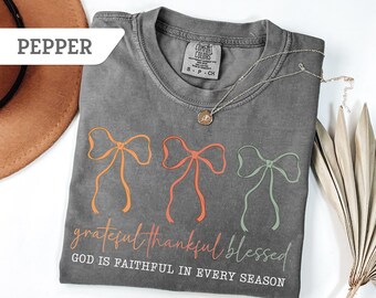 May include: A gray t-shirt with three bow designs in orange, coral, and green. The text "grateful thankful blessed" and "GOD IS FAITHFUL IN EVERY SEASON" are printed below the bows. A silver necklace with a pendant is draped across the shirt.