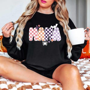 May include: Black sweatshirt with a checkered "Mama" graphic in orange, purple, and white with Halloween-themed accents like stars, bats, and a spider.