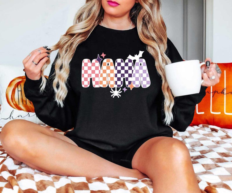 May include: Black sweatshirt with a checkered "Mama" graphic in orange, purple, and white with Halloween-themed accents like stars, bats, and a spider.