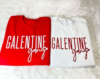 May include: Two sweatshirts, one red and one white, with the text "Galentine gang" printed on them. The text on the red sweatshirt is white, and the text on the white sweatshirt is red.