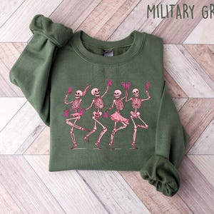 Pode incluir: Uma camisa de moletom verde com um gr&aacute;fico de quatro esqueletos vestindo tutus rosa e segurando fitas rosa. Os esqueletos est&atilde;o dan&ccedil;ando e sorrindo. O texto "MILITARY GREEN" est&aacute; impresso em branco no topo da imagem.