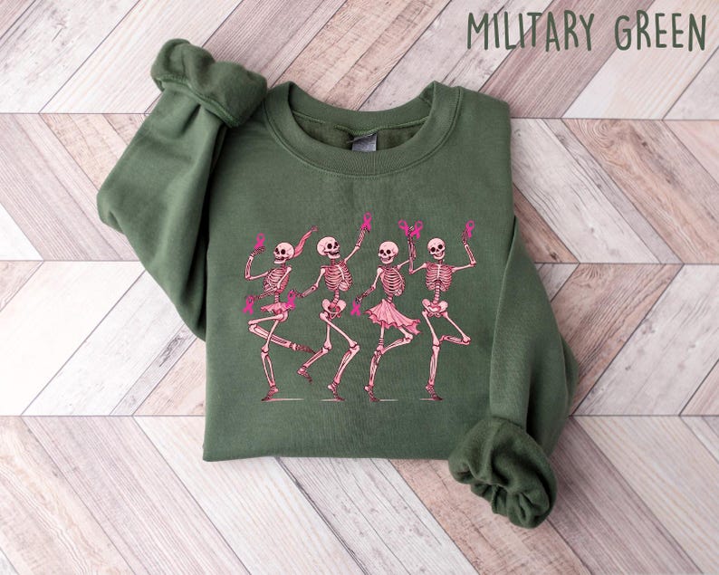Pode incluir: Uma camisa de moletom verde com um gr&aacute;fico de quatro esqueletos vestindo tutus rosa e segurando fitas rosa. Os esqueletos est&atilde;o dan&ccedil;ando e sorrindo. O texto "MILITARY GREEN" est&aacute; impresso em branco no topo da imagem.