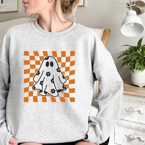 May include: A grey sweatshirt featuring a graphic of a ghost with black eyes and floral details on a checkered orange and white background.
