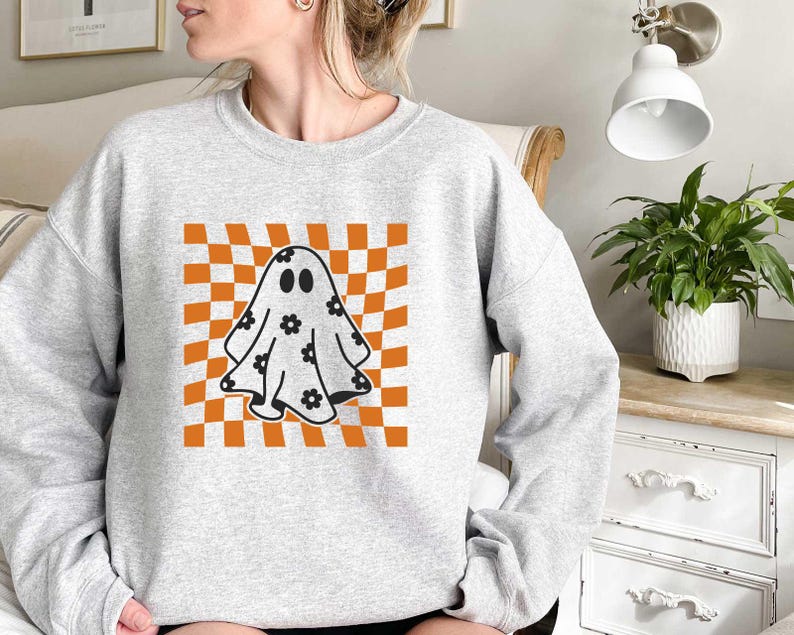 May include: A grey sweatshirt featuring a graphic of a ghost with black eyes and floral details on a checkered orange and white background.