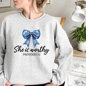 Może przedstawiać: Szara bluza z niebieską wstążką z dżinsu i nadrukiem "She is worthy Proverbs 31" z przodu.