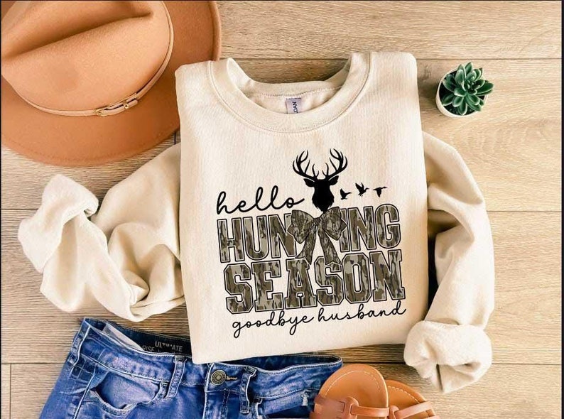 May include: A cream-colored sweatshirt with the text "hello hunting season goodbye husband" in black lettering. The text is surrounded by a camouflage pattern with a silhouette of a deer with antlers and a bow.