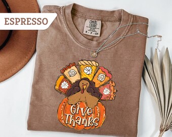 May include: Espresso-colored t-shirt with a Thanksgiving-themed graphic. The design features a turkey, pumpkin, and slices of pie, with the words "Give Thanks" on the pumpkin. A silver necklace is draped across the shirt.