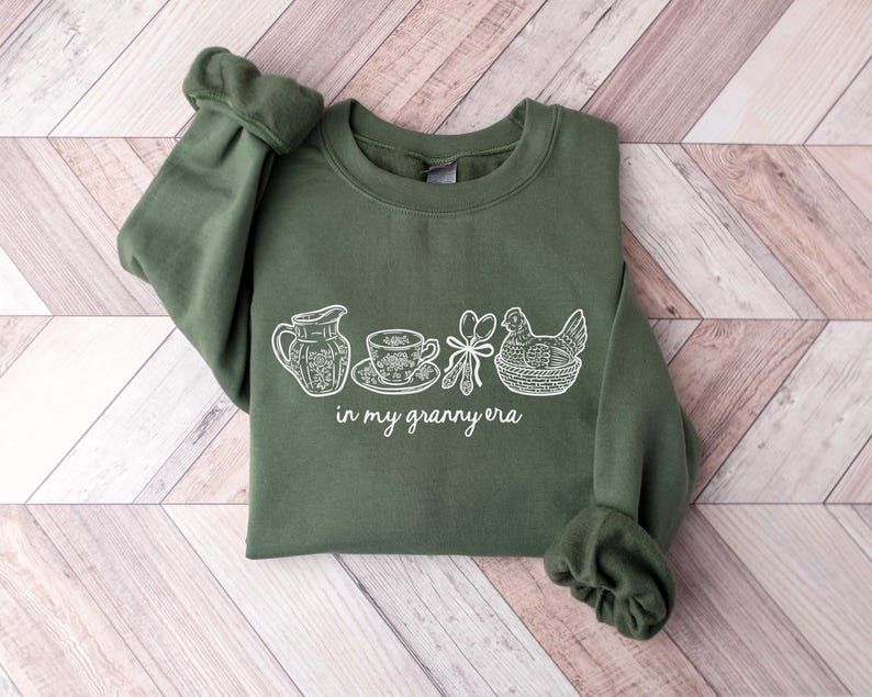 Puede incluir: Una sudadera verde oliva con un dibujo lineal blanco de una jarra, una taza de t&eacute;, utensilios y una gallina en una cesta. El texto "in my granny era" est&aacute; debajo. Las mangas est&aacute;n dobladas.