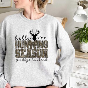 May include: A gray sweatshirt with a camouflage pattern and the text "hello hunting season goodbye husband". The design features a deer with antlers and a bow.