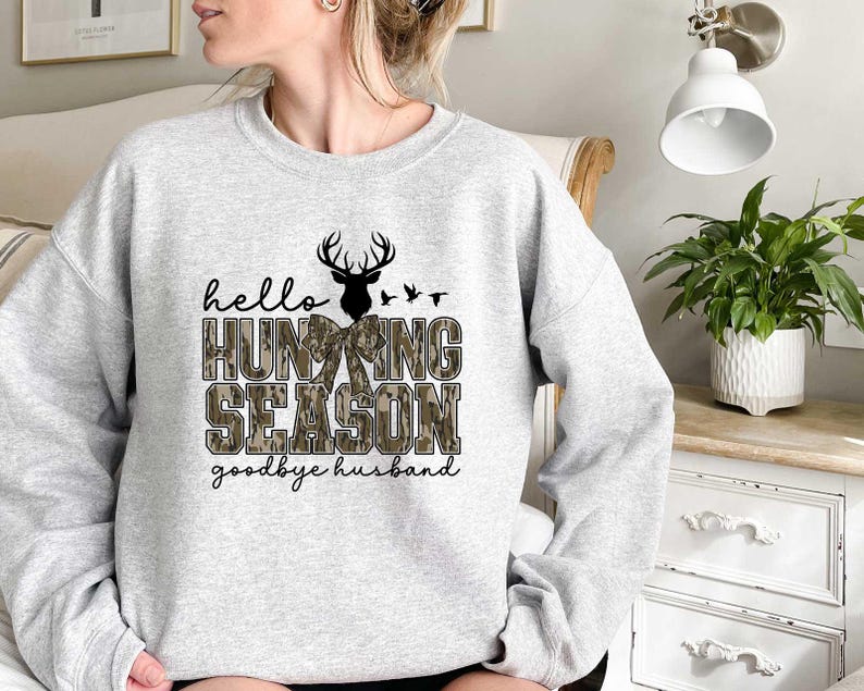 May include: A gray sweatshirt with a camouflage pattern and the text "hello hunting season goodbye husband". The design features a deer with antlers and a bow.