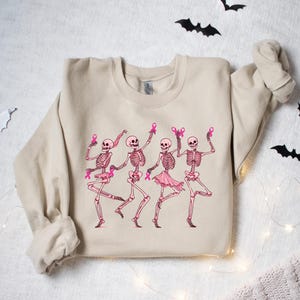 Pode incluir: Uma camisa de moletom bege com um gr&aacute;fico de quatro esqueletos usando fitas rosa e dan&ccedil;ando. Os esqueletos s&atilde;o rosa e branco.