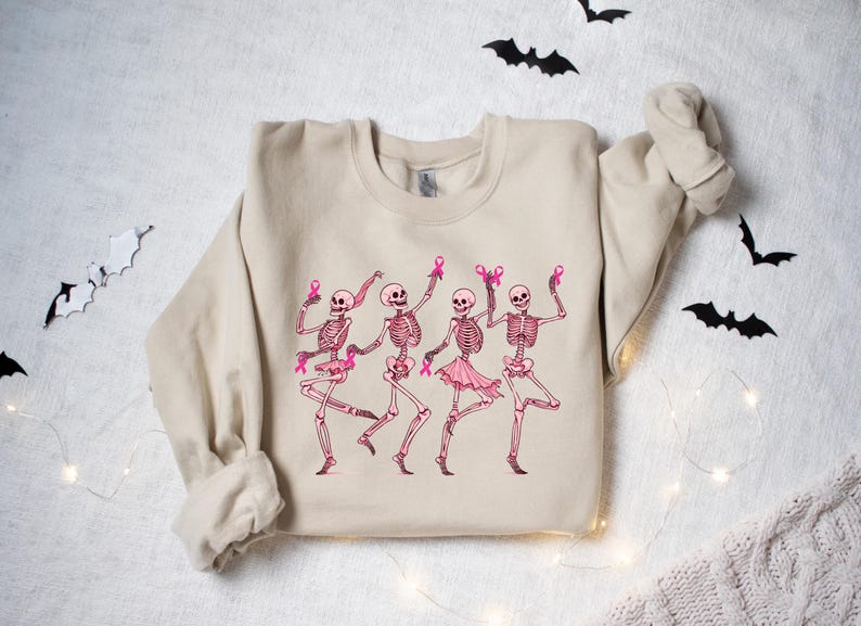 Pode incluir: Uma camisa de moletom bege com um gr&aacute;fico de quatro esqueletos usando fitas rosa e dan&ccedil;ando. Os esqueletos s&atilde;o rosa e branco.